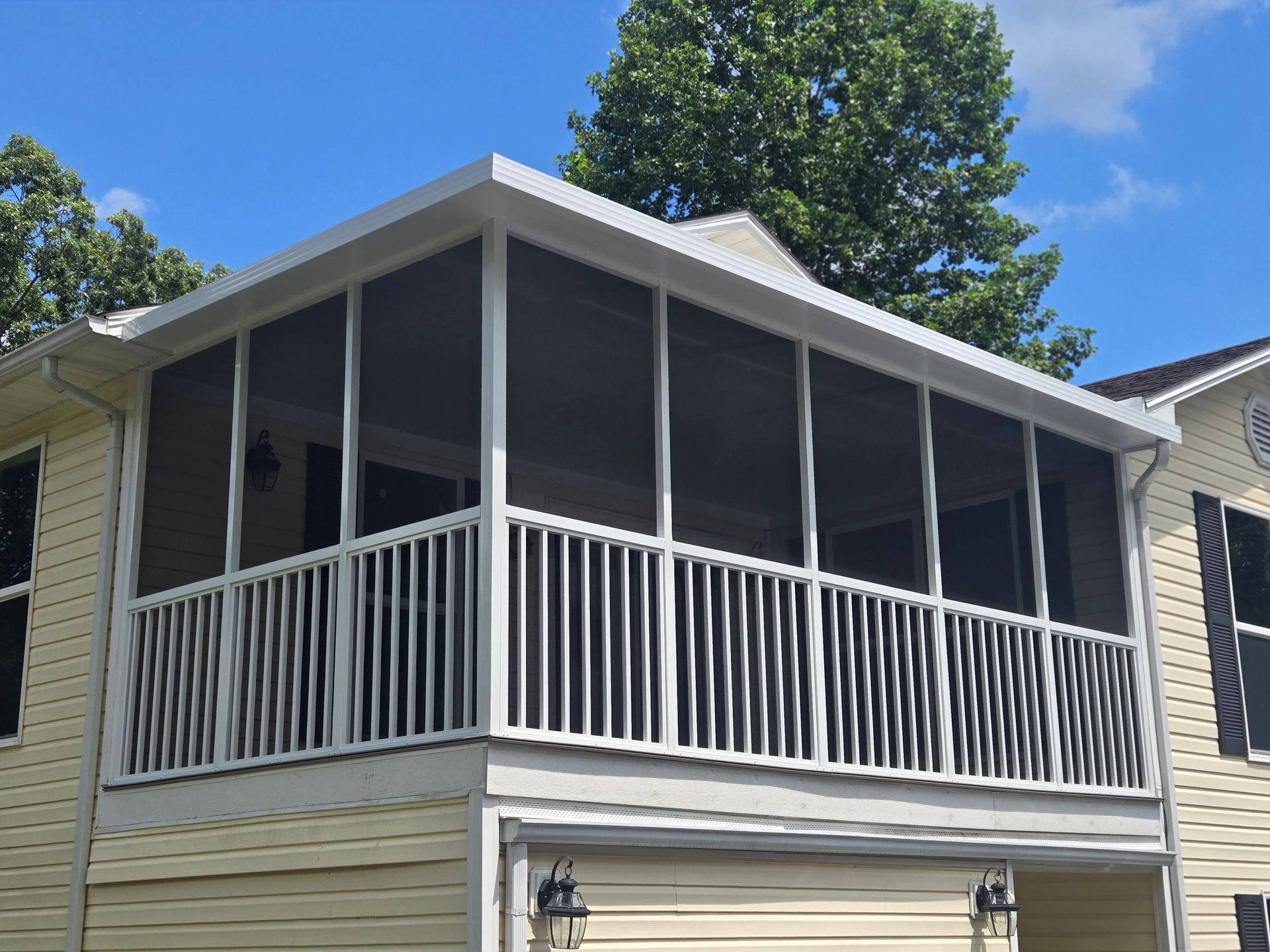 A second-story screened-in porch with white railings, attached to the side of a yellow-sided house under a blue sky.