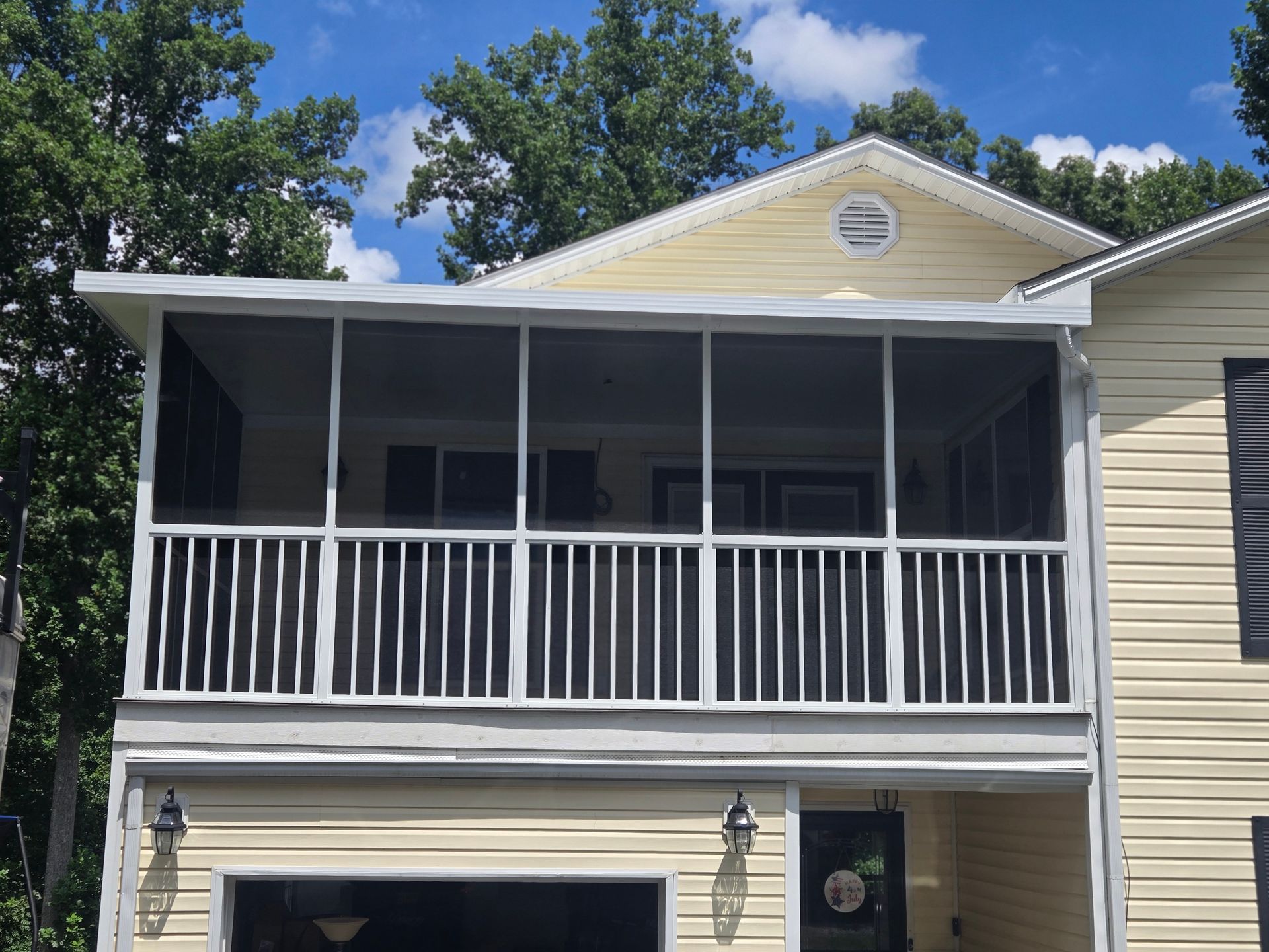 A yellow house exterior featuring a second-story screened-in porch with white railings, set against a backdrop of trees.