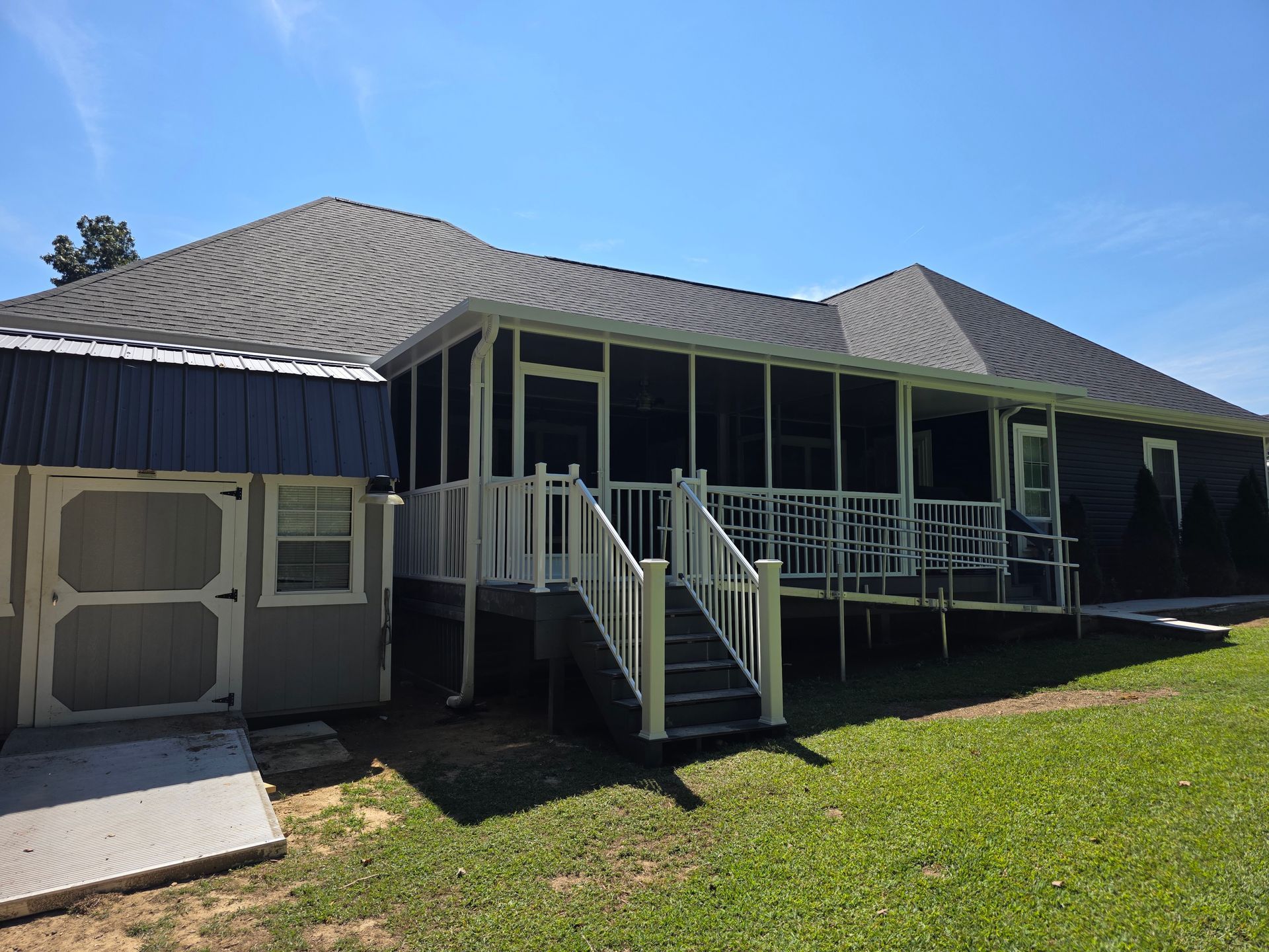 A dark-sided house with a shingled roof, a screened-in porch with white railings, and a small attached shed.
