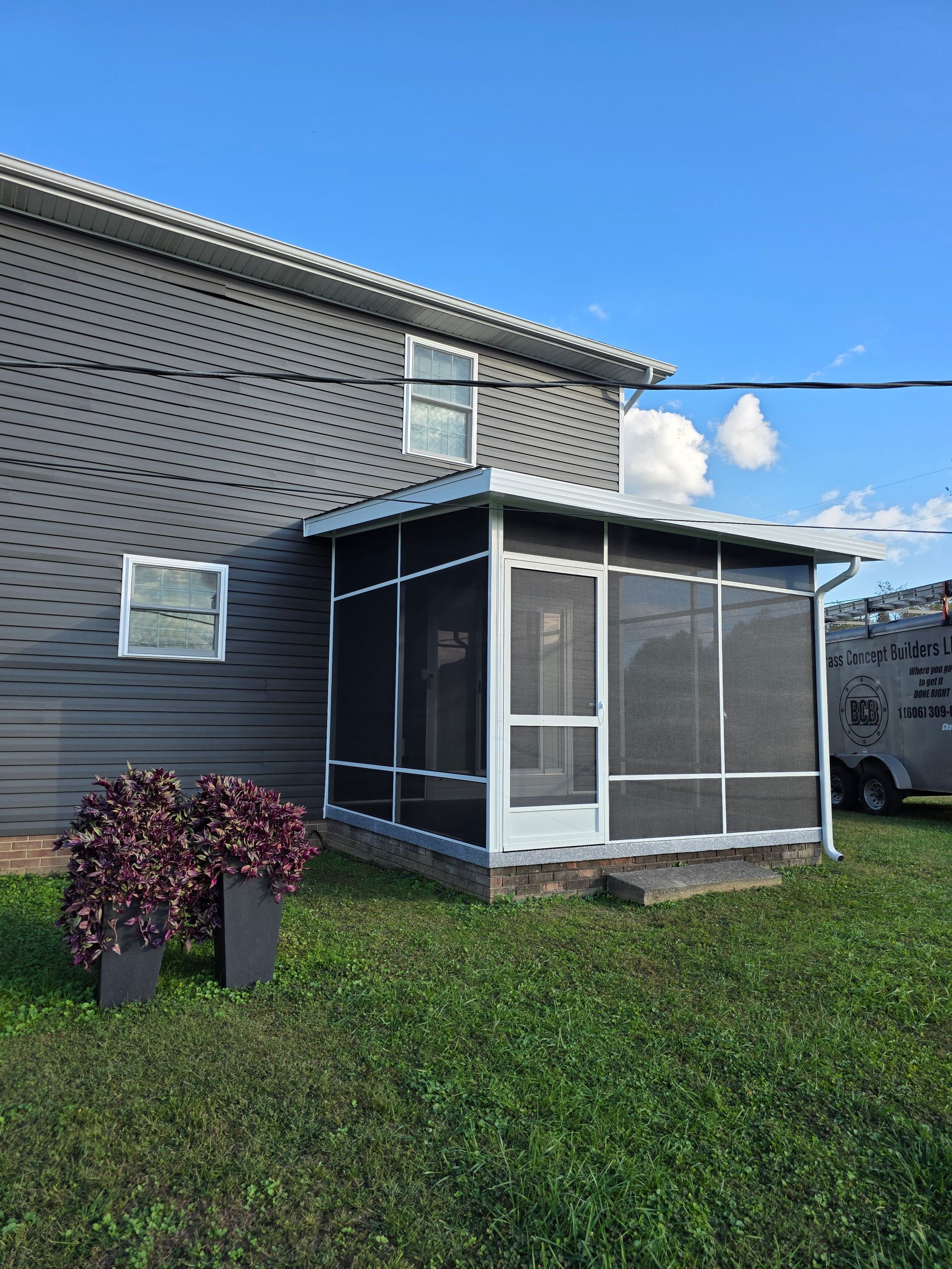 A gray house with a white screened-in porch and two large, dark planters of purple foliage in the grass.