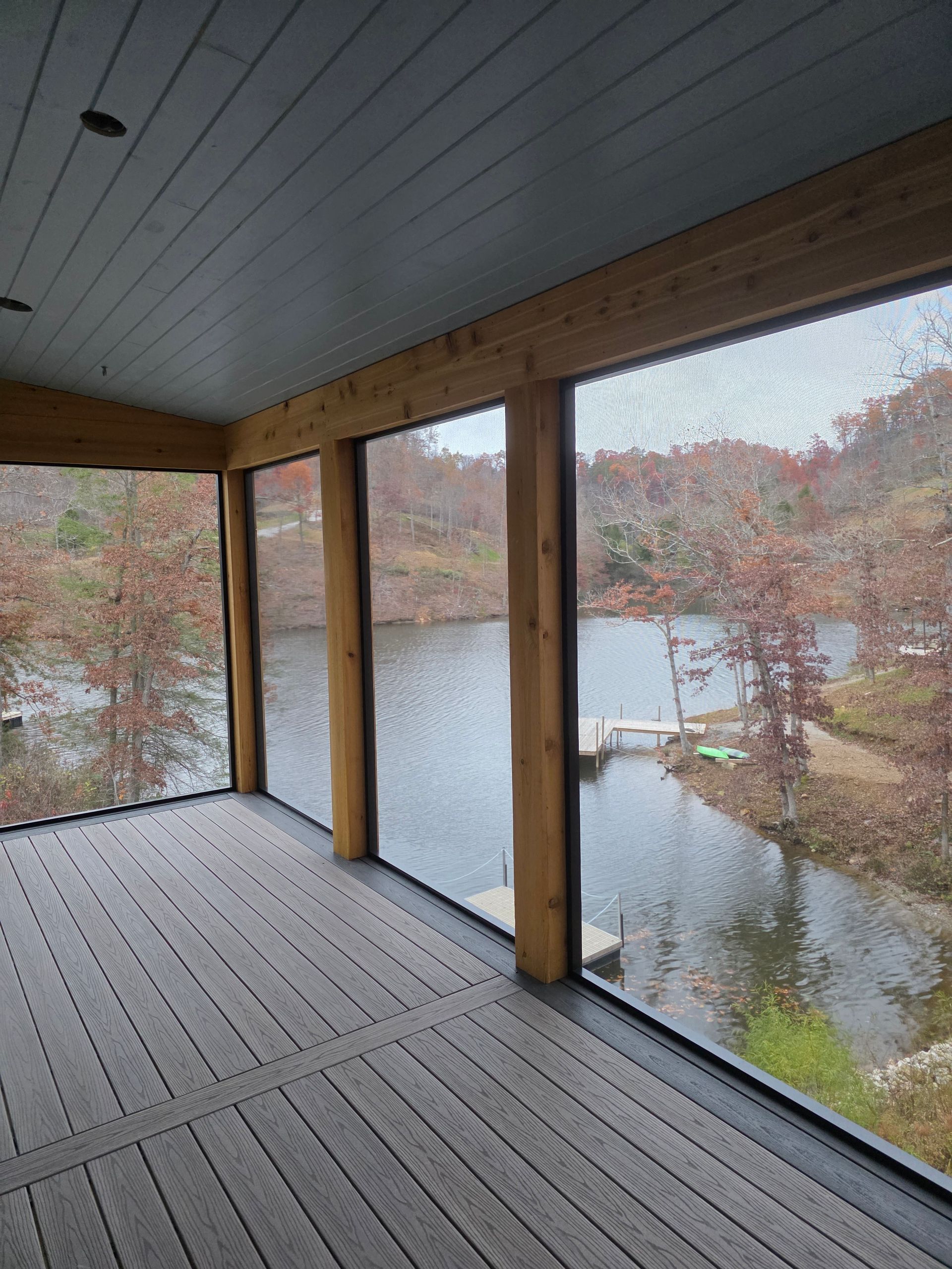 A screened-in porch with wood posts and gray composite decking overlooking a lake with autumn trees.