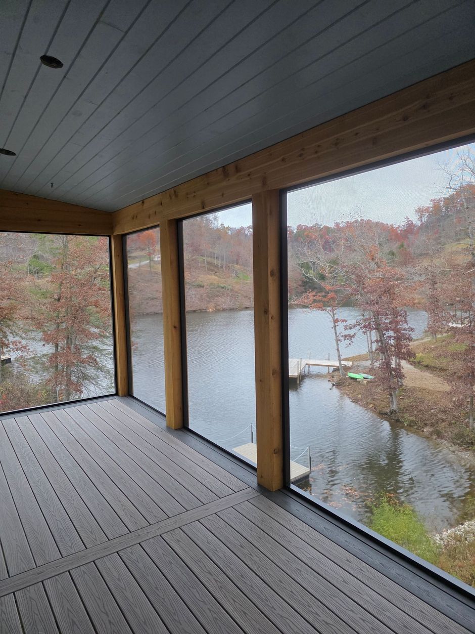 A screened-in porch with wood posts and gray composite decking overlooking a lake with autumn trees and a small dock.