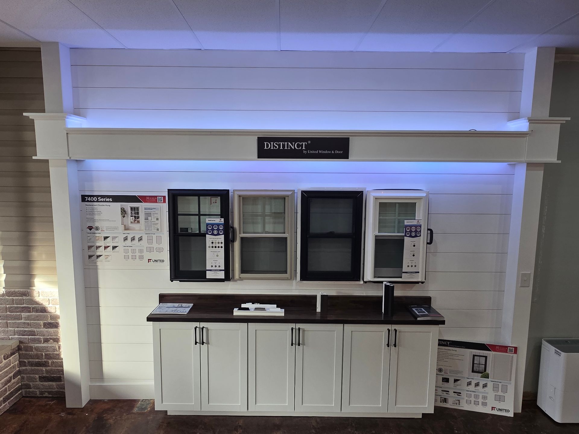 A showroom display featuring four sample windows mounted above white cabinets against a white shiplap wall with lighting.