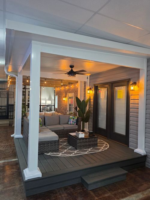 A covered outdoor deck with gray wooden floor, wicker furniture, a round rug, ceiling fan, and string lights at night.