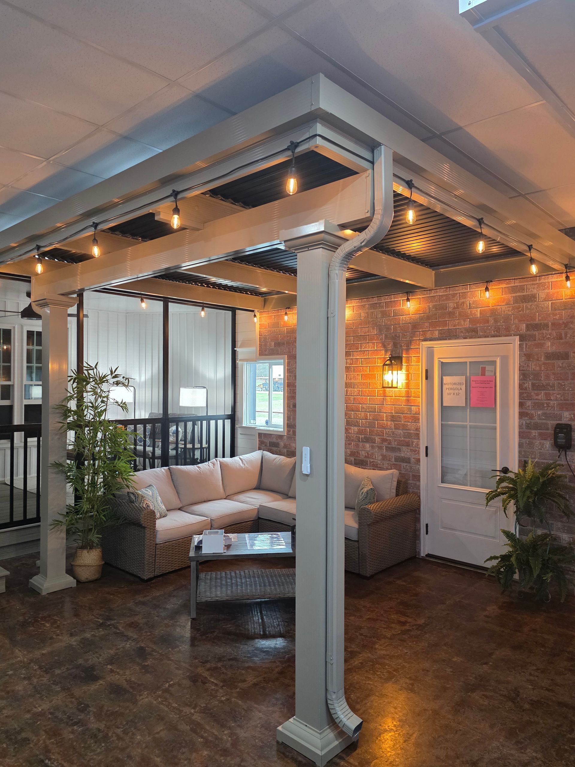 A cozy covered patio with a sectional sofa, a coffee table, string lights, and a white door against a brick wall.