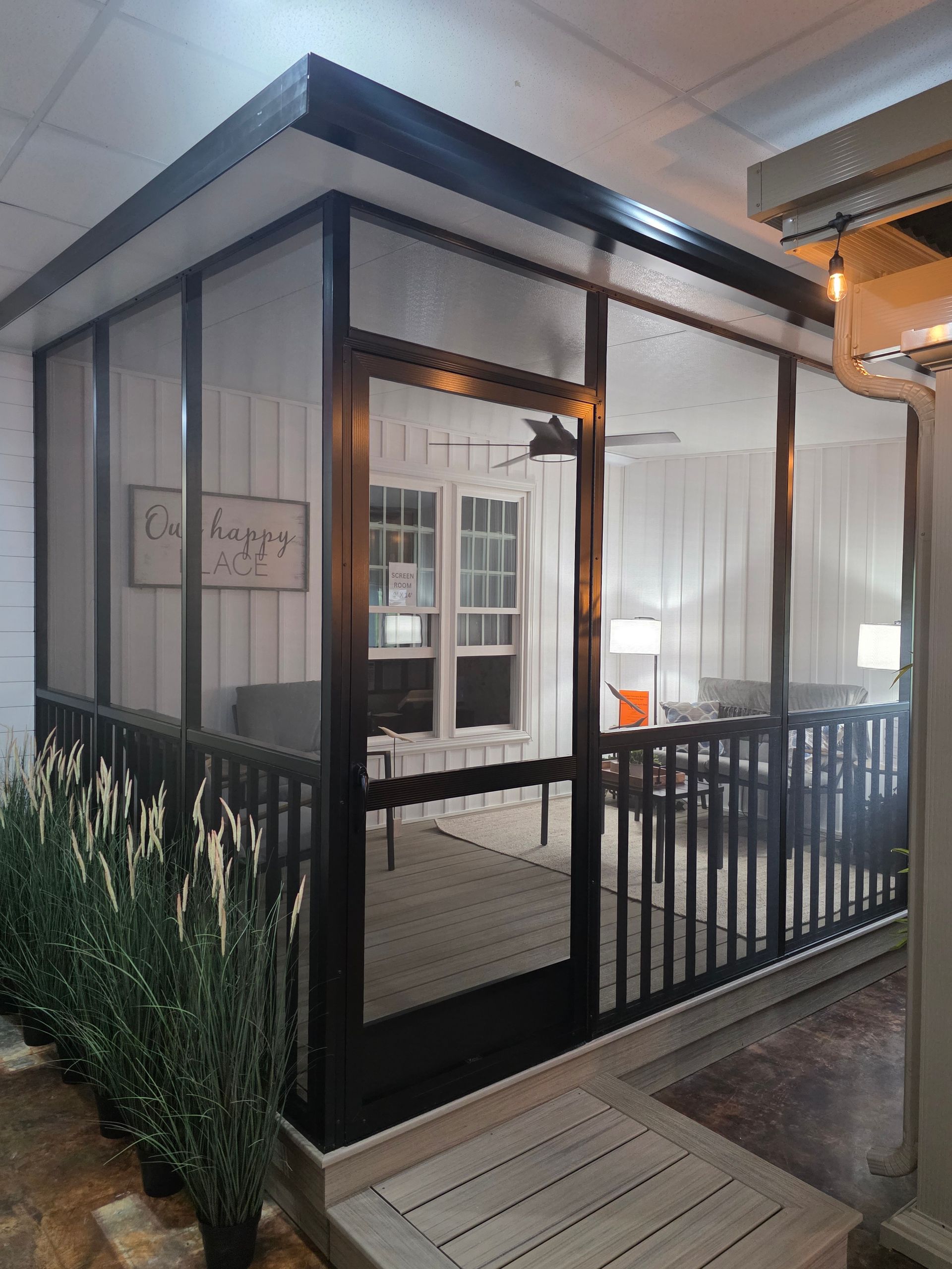 A screened-in porch with a dark frame, a door, and wooden decking, featuring potted plants in the foreground.