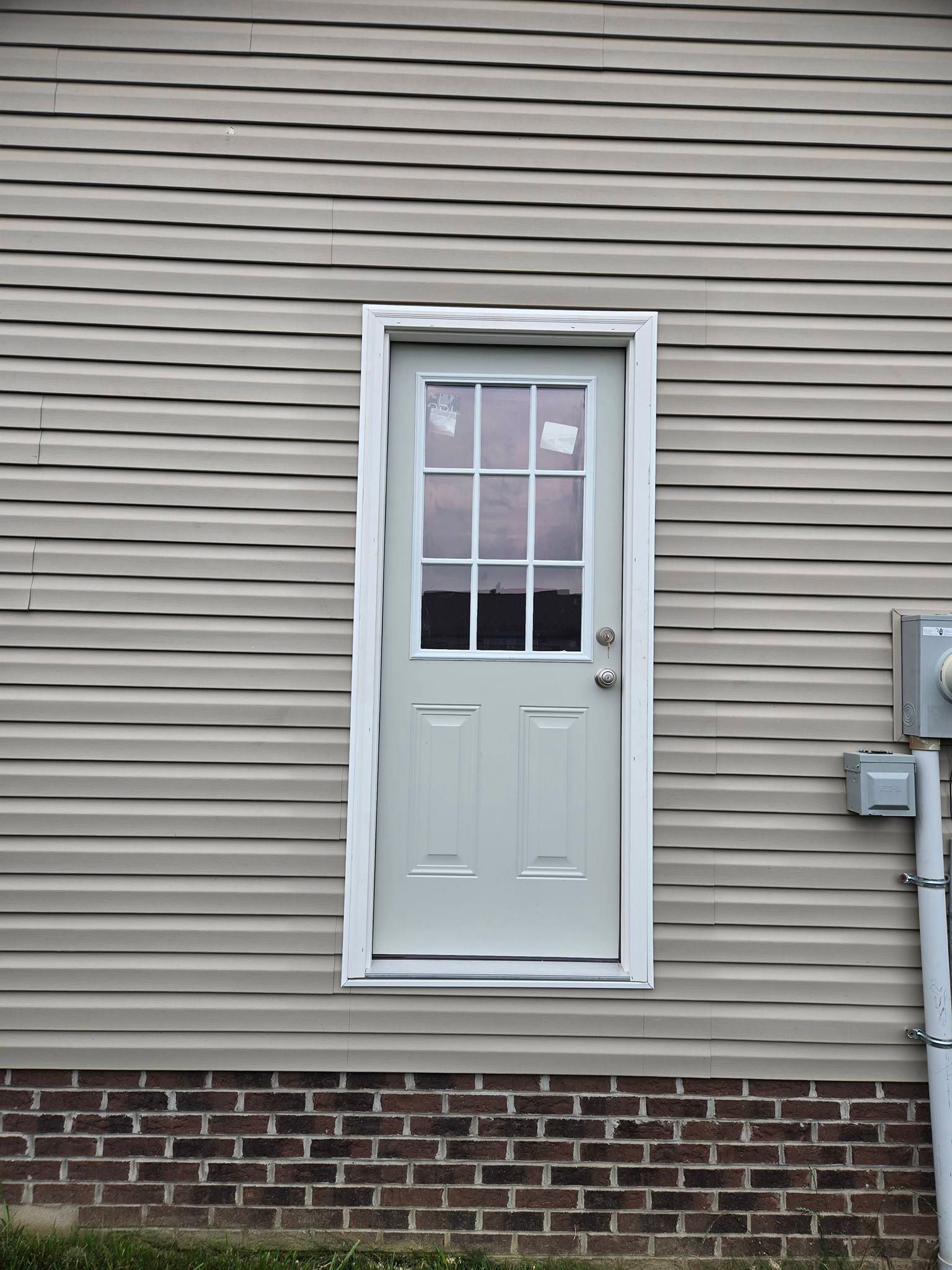 A light-colored, narrow exterior door with a glass window above two panels, set into a beige vinyl-sided wall above brick.
