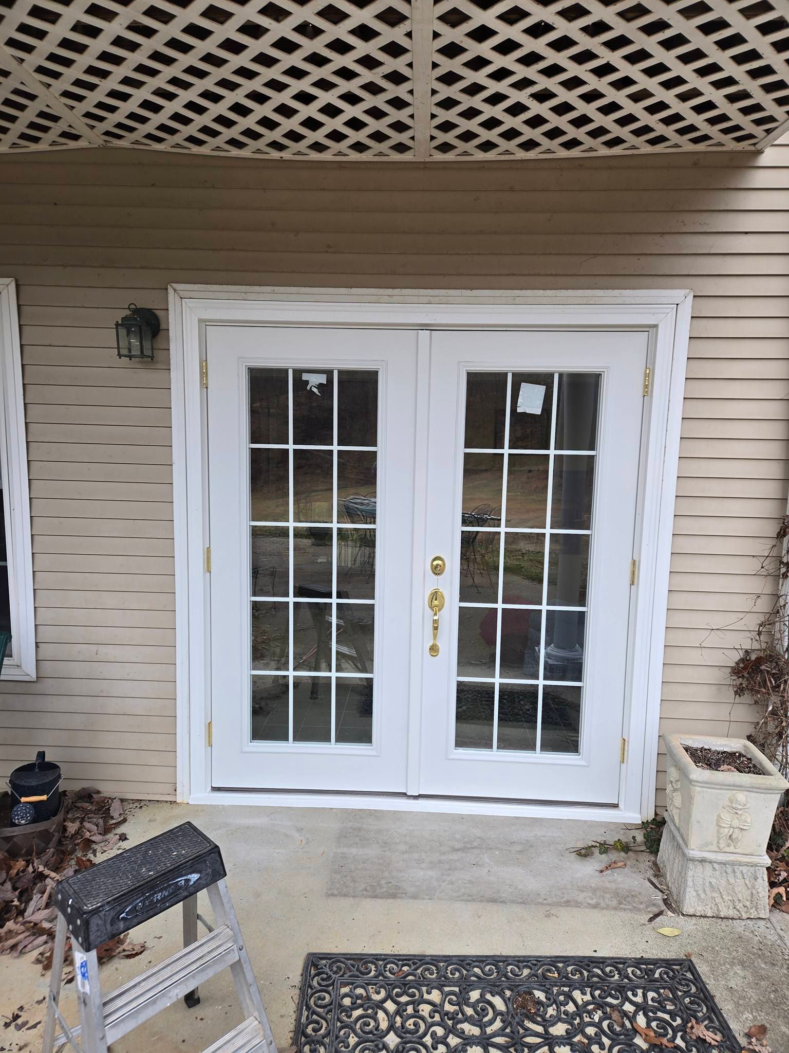White double French doors with glass panes installed in a tan-sided wall, with a small ladder nearby on a concrete porch.