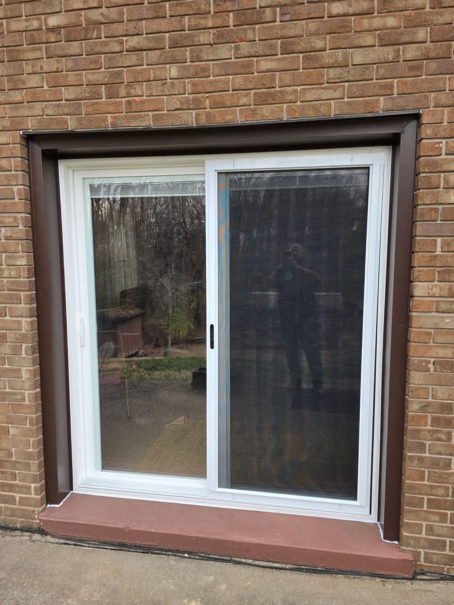 A white sliding glass door with a screen installed in a brown brick exterior wall, set above a raised red concrete step.