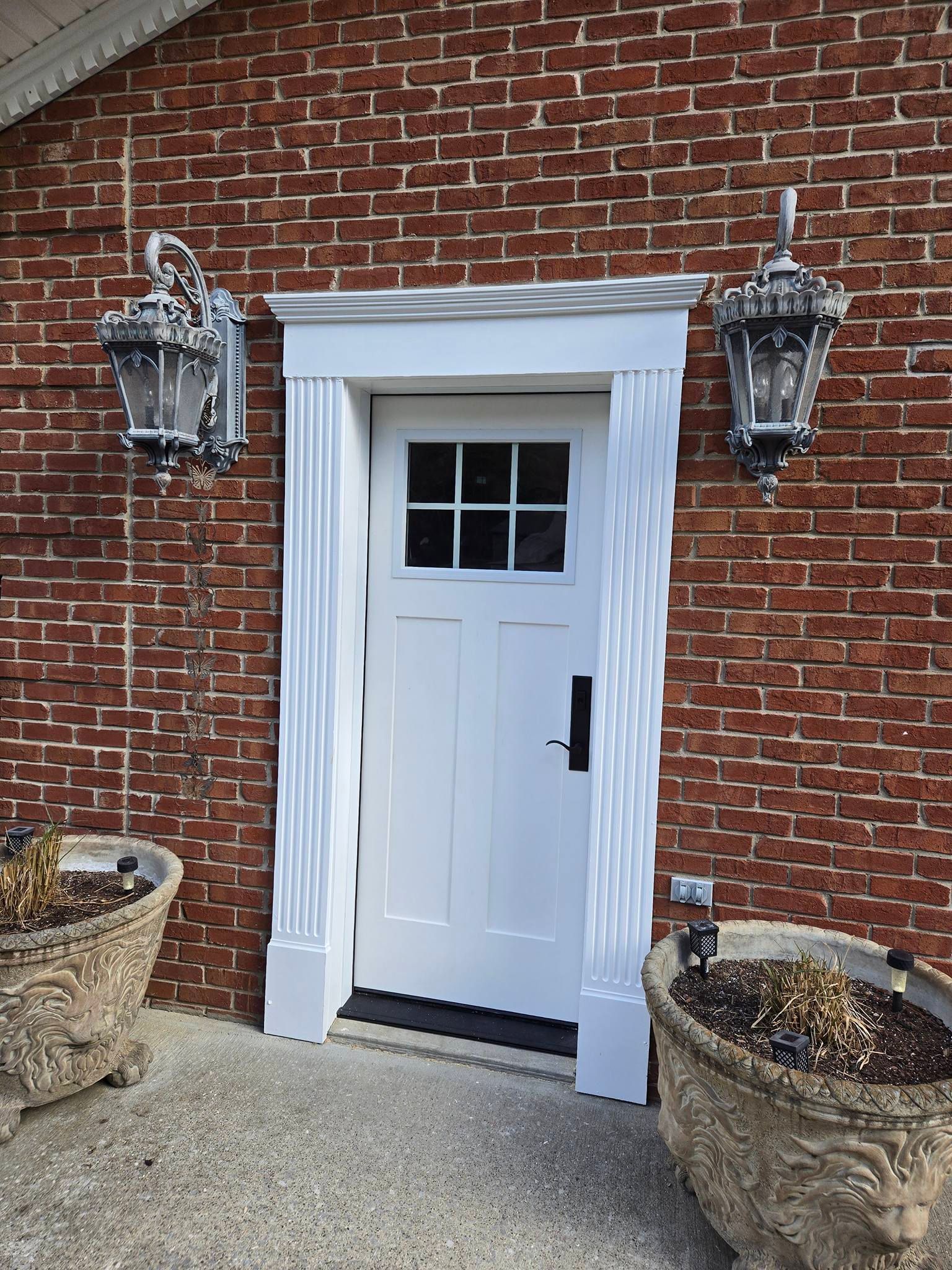 A white door with a six-pane window set in a brick wall, framed by white fluted columns and flanked by outdoor lanterns.