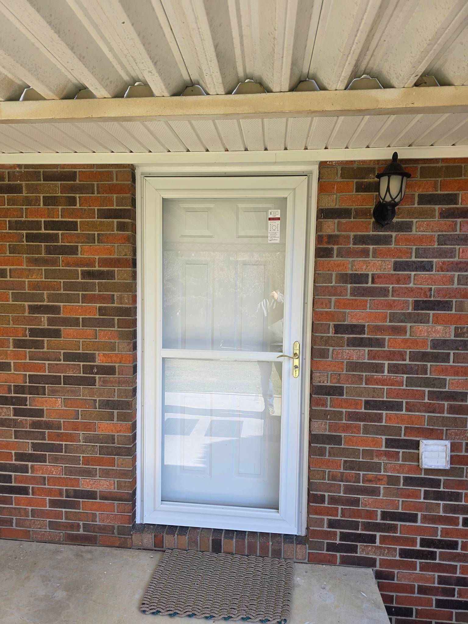 A white storm door installed on a multi-toned red brick house exterior with a small welcome mat on the concrete porch.