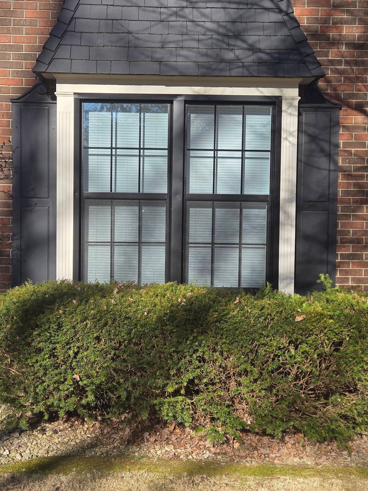 A brick house exterior featuring a double-pane window with black shutters and a small, shingled roof overhang above.