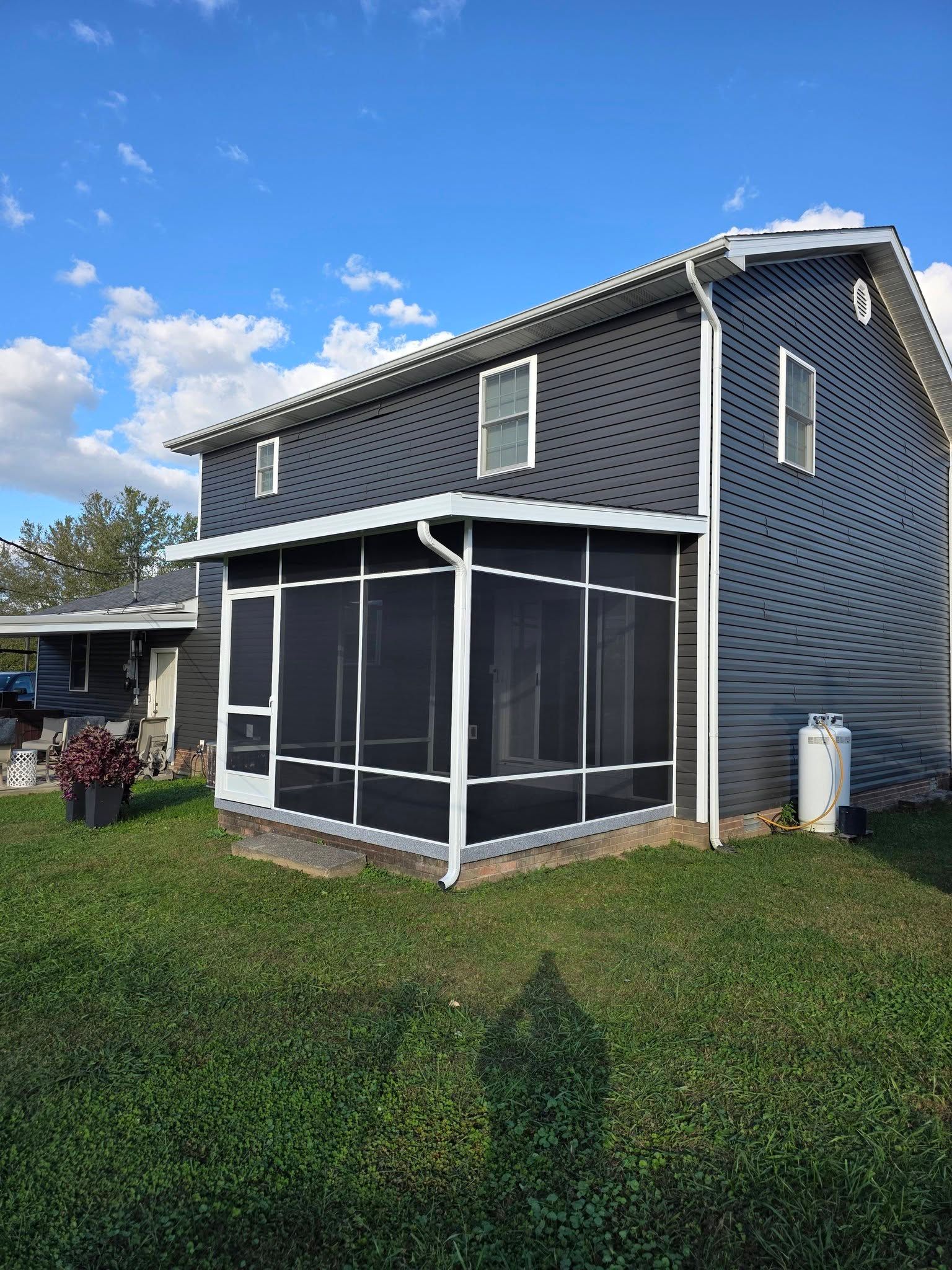 A two-story house with dark gray horizontal siding and a white-framed screened porch in a grassy yard under a blue sky.