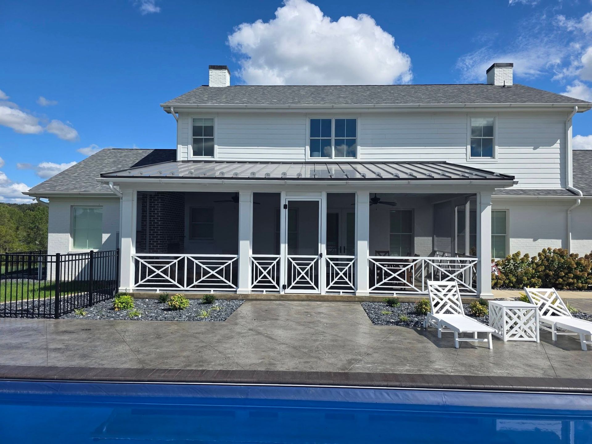 A white two-story house with a screened porch, a gray patio, pool, and two lounge chairs under a blue sky.