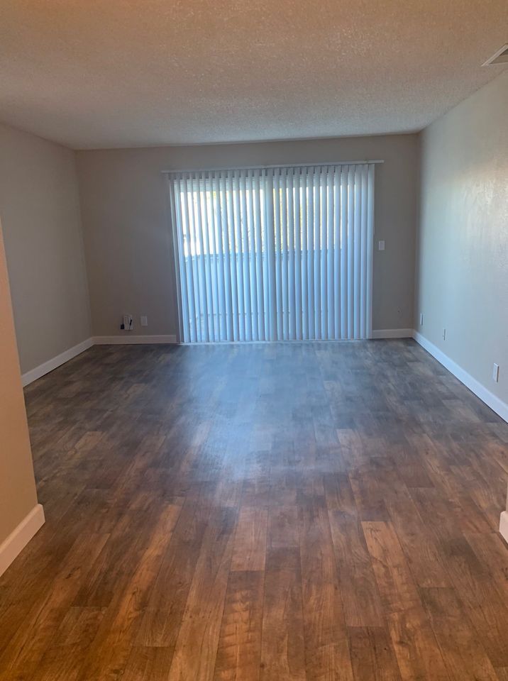 Empty living room with dark wood floors and vertical blinds on the back window.