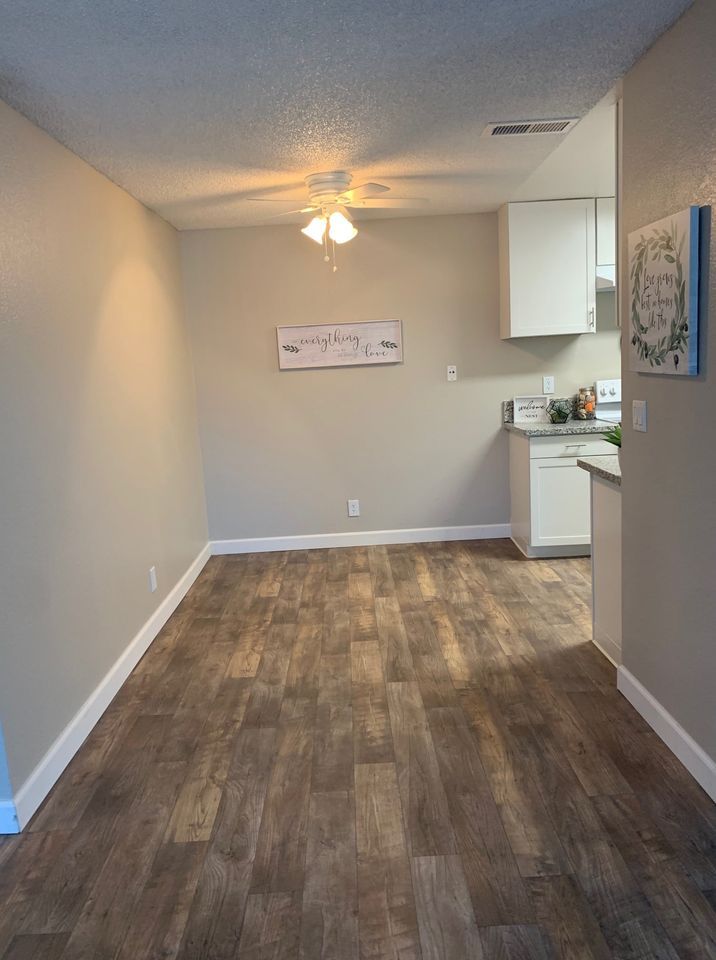 Empty room with wood flooring, beige walls, ceiling fan, and a small kitchen area on the right