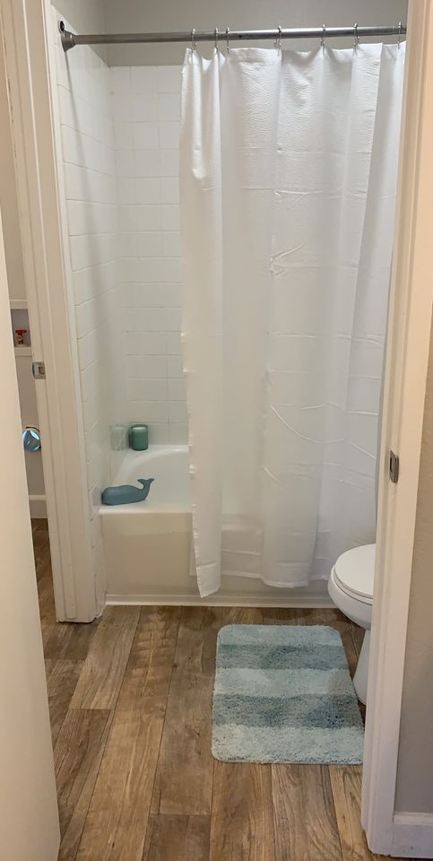 Small bathroom with shower curtain, teal accessories, toilet, and blue bath mats on wood-look floor.