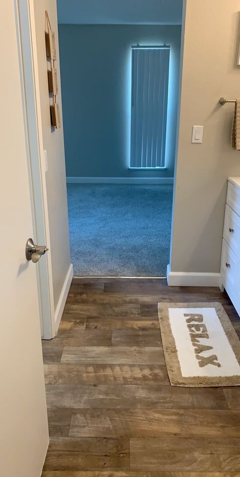 Apartment hallway with wood-look floor, blue carpet room ahead, and a white welcome mat by the door