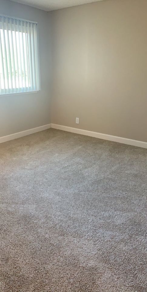 Empty beige carpeted room with white walls, a window with blinds, and a power outlet.