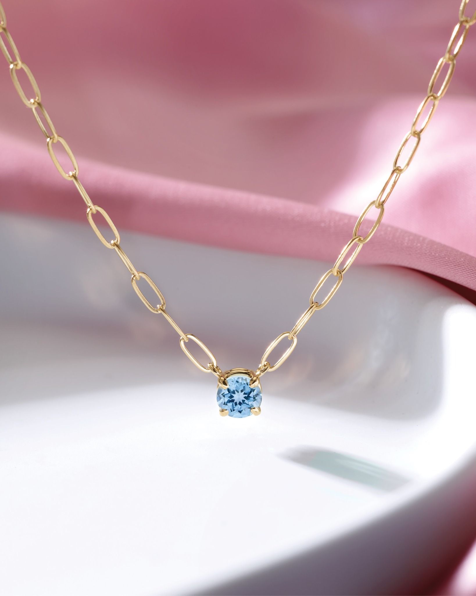 Gold necklace with a light blue gemstone, on a pink and white background.