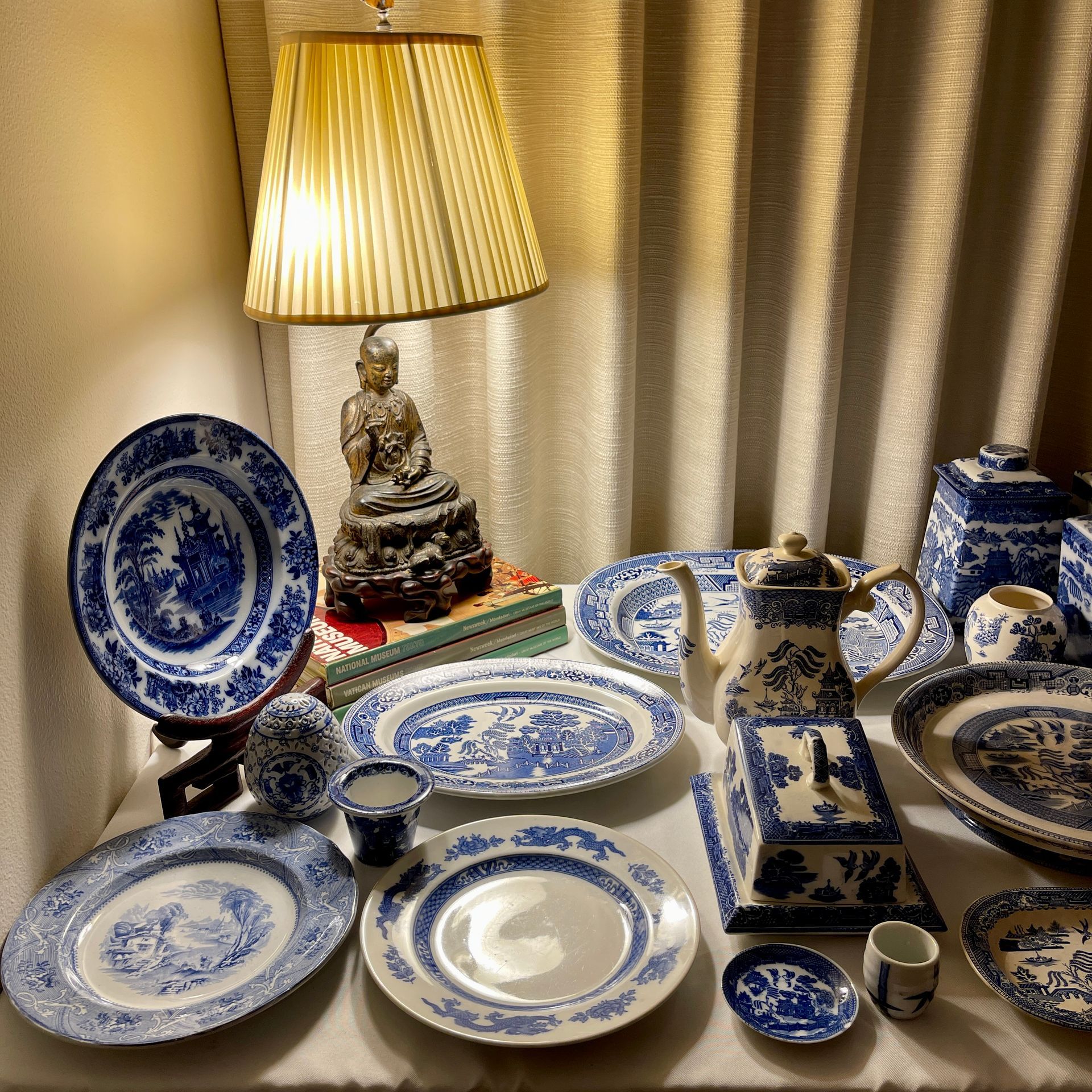 Blue and white porcelain plates, teapot, and cups displayed on a table next to a lamp and small statue.