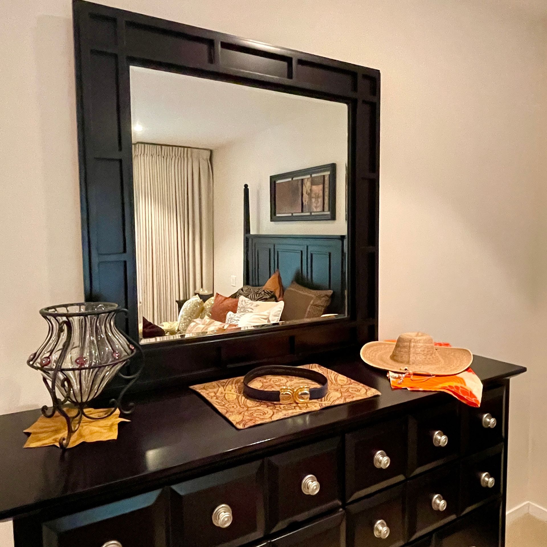 Black dresser with mirror, decorated with a lamp, belt, hat, and decorative cloth. Reflection shows bed and curtains.