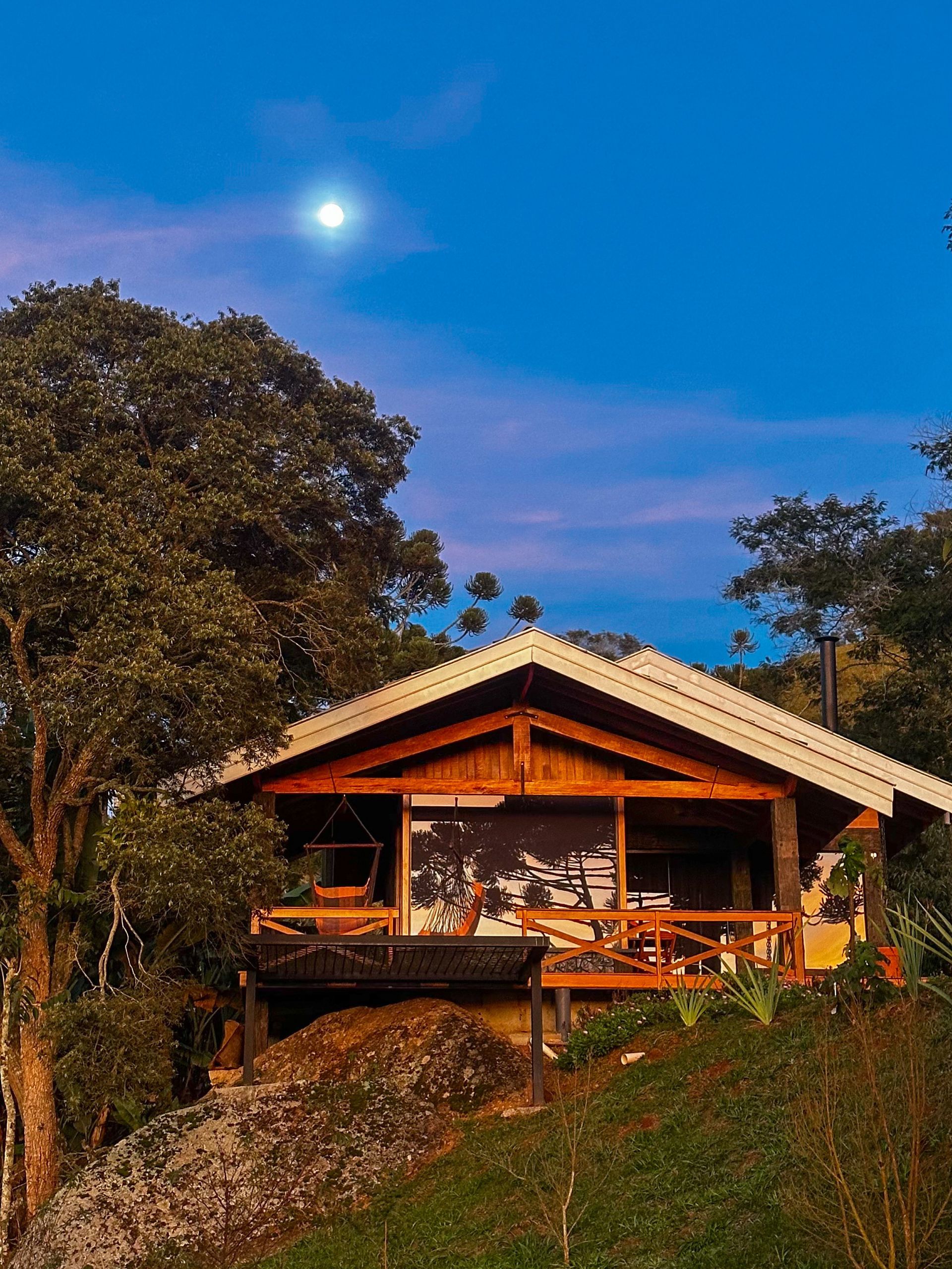Cabana de madeira numa colina ao entardecer, emoldurada por árvores, com uma lua brilhante num céu azul.