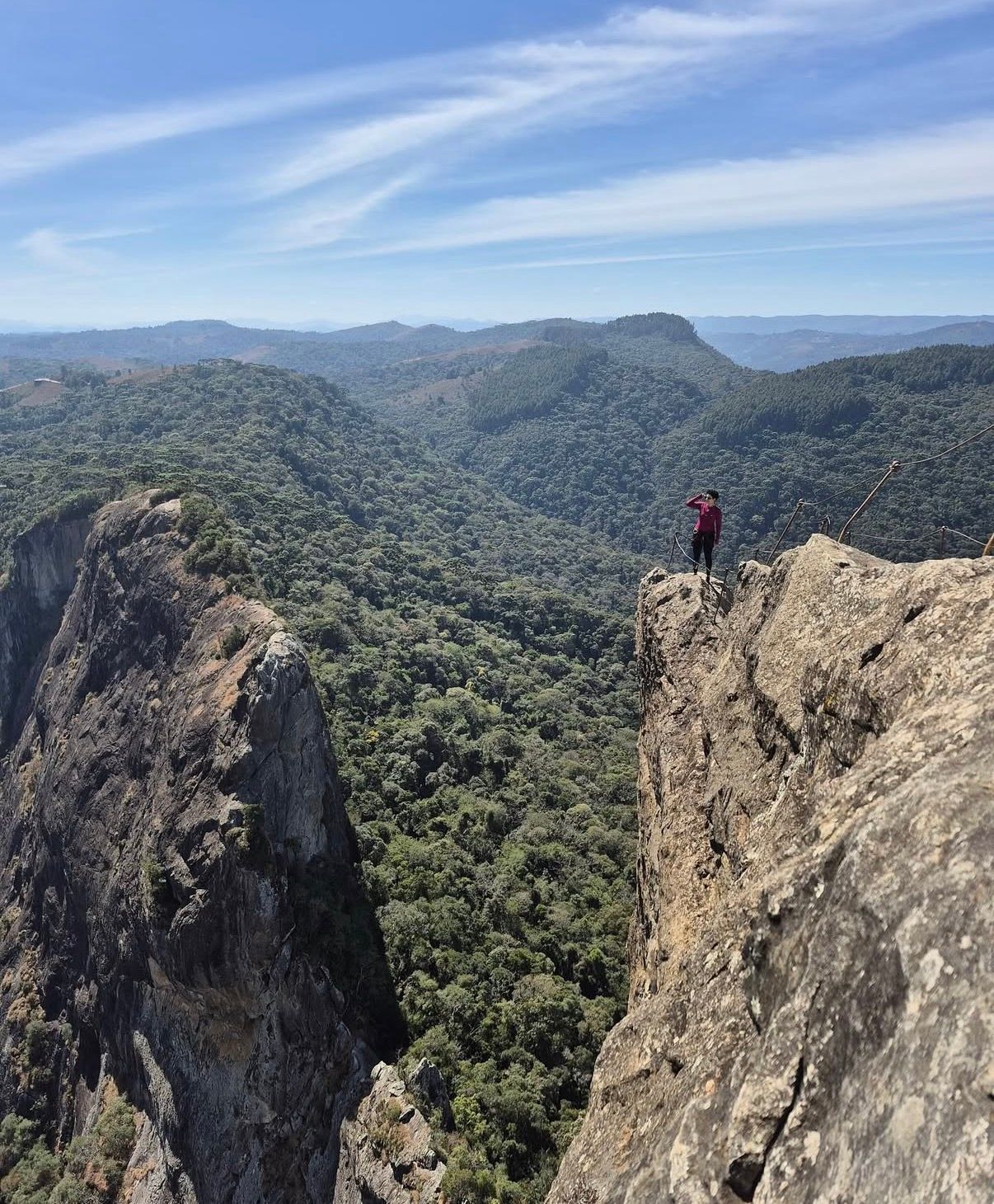 Pessoa em pé em um penhasco rochoso com vista para um vale arborizado e cristas azuis distantes.