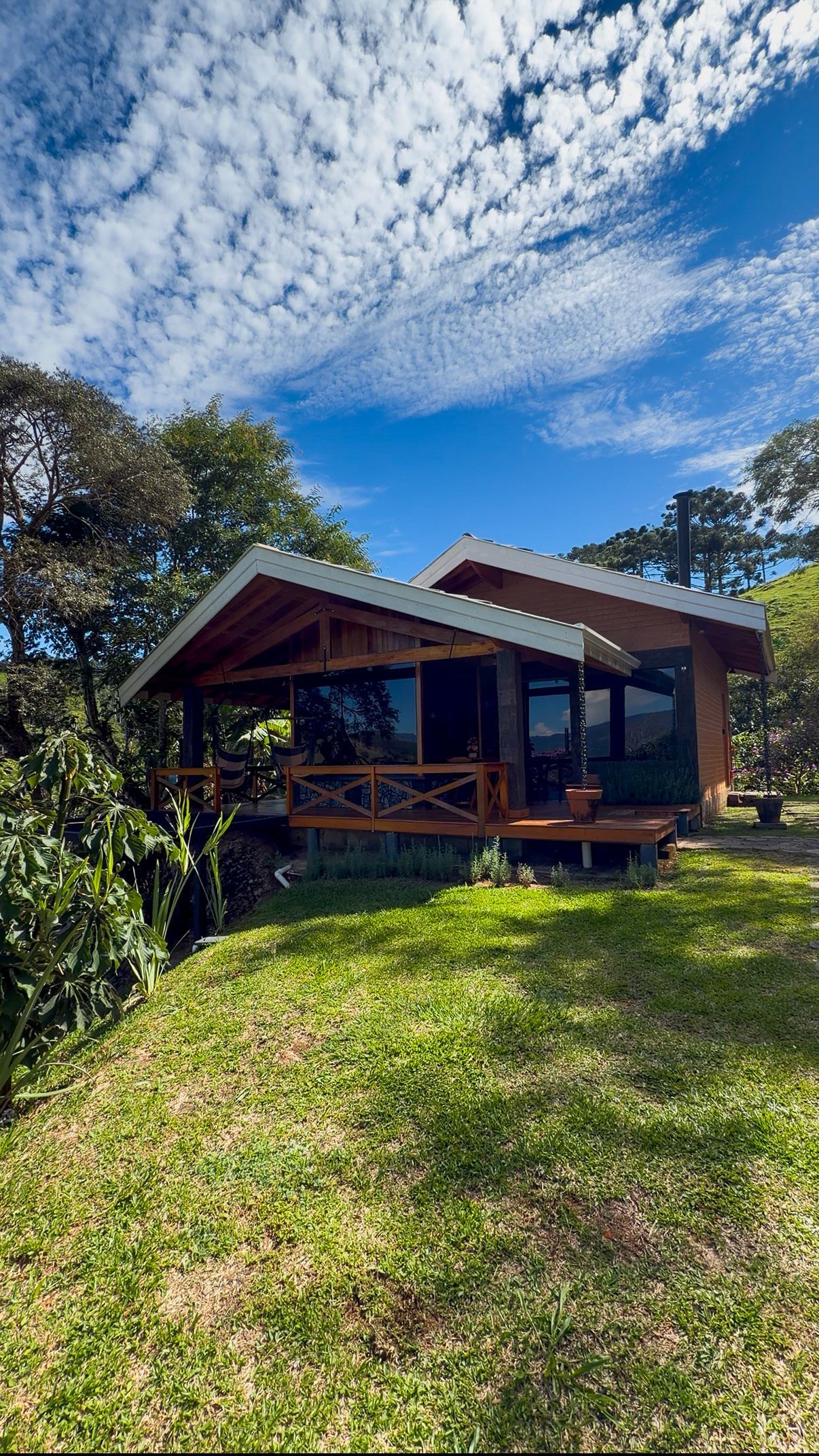Cabana rústica com varanda em uma clareira gramada e ensolarada sob um céu azul com nuvens esparsas.