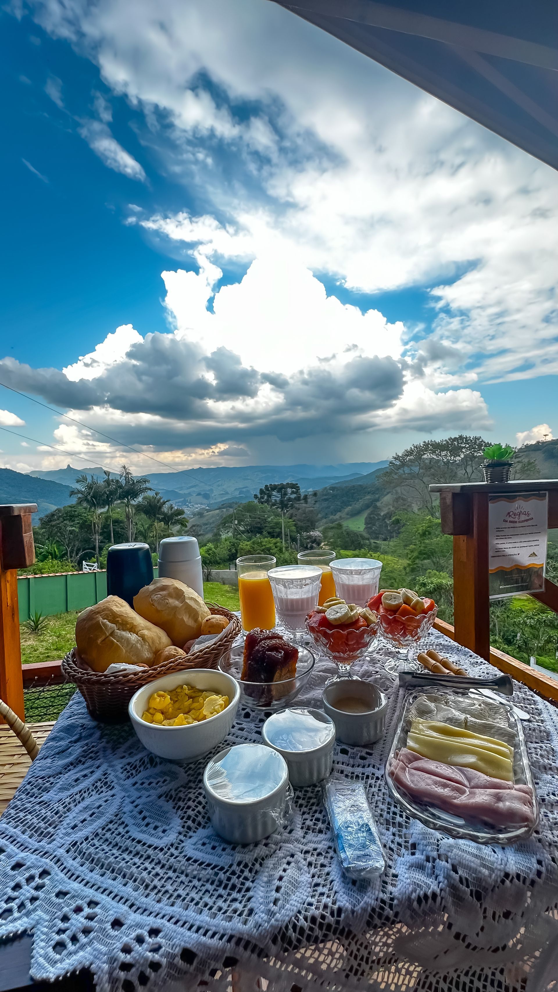 Mesa de café da manhã ao ar livre com doces, frutas e bebidas com vista para uma montanha encoberta por nuvens.