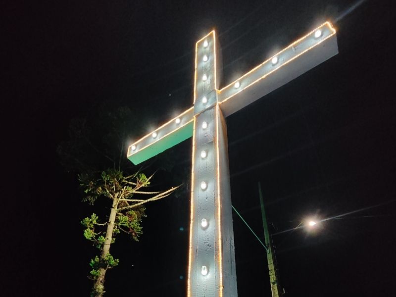 Cruz iluminada à beira da estrada à noite, contra um céu escuro, com uma pequena árvore ao lado.
