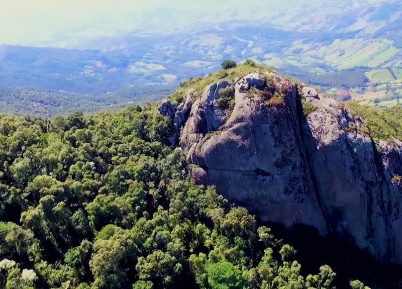 Pico rochoso elevando-se acima de uma densa floresta verde sob um céu azul enevoado.