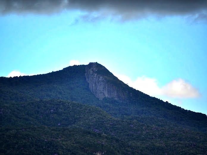 Céu azul sobre um pico de montanha arborizado com uma face rochosa íngreme.