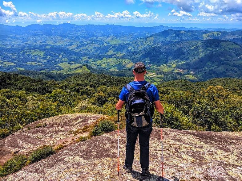 Caminhante com mochila e bastões de caminhada contemplando um vale montanhoso exuberante sob um céu azul.