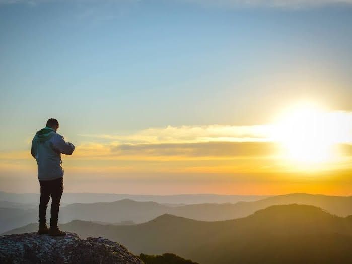 Pessoa em pé em um pico rochoso ao nascer do sol, contemplando montanhas em camadas e um céu dourado brilhante.