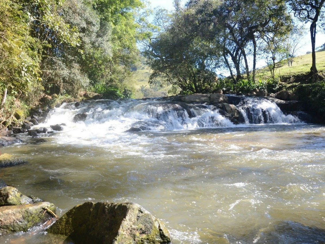 Pequena cachoeira de rio caindo sobre rochas em uma área arborizada e ensolarada.