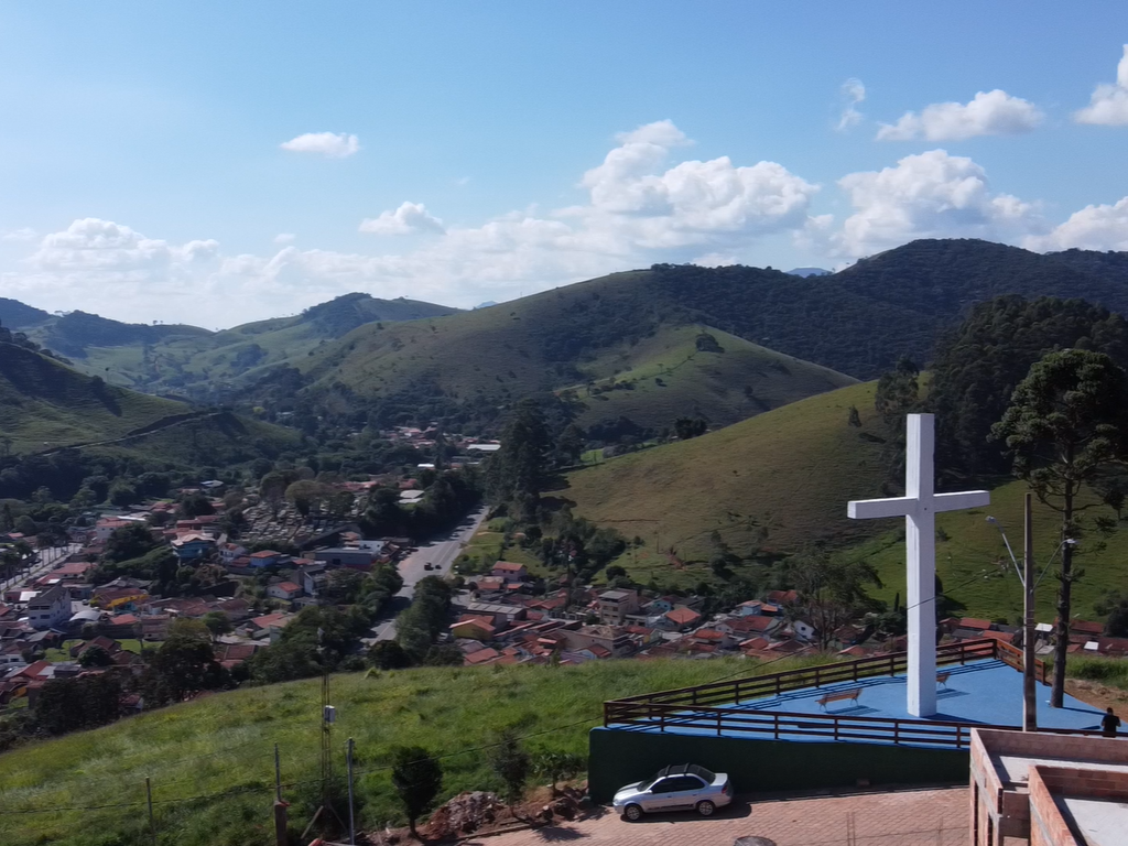 Vista do alto de uma colina de uma cidade e montanhas verdes, com uma grande cruz branca em primeiro plano.
