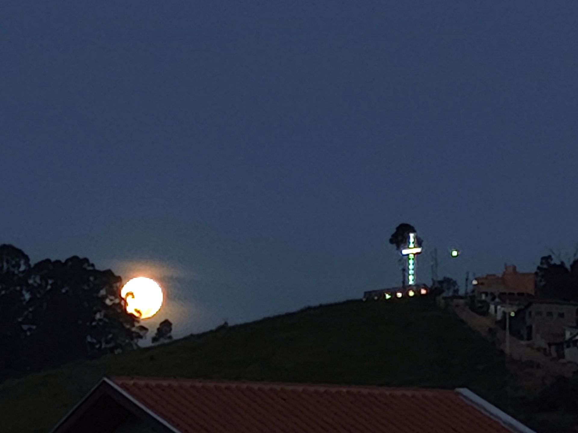 Horizonte ao entardecer com lua brilhante, árvores escuras, uma colina gramada e uma torre iluminada à direita.