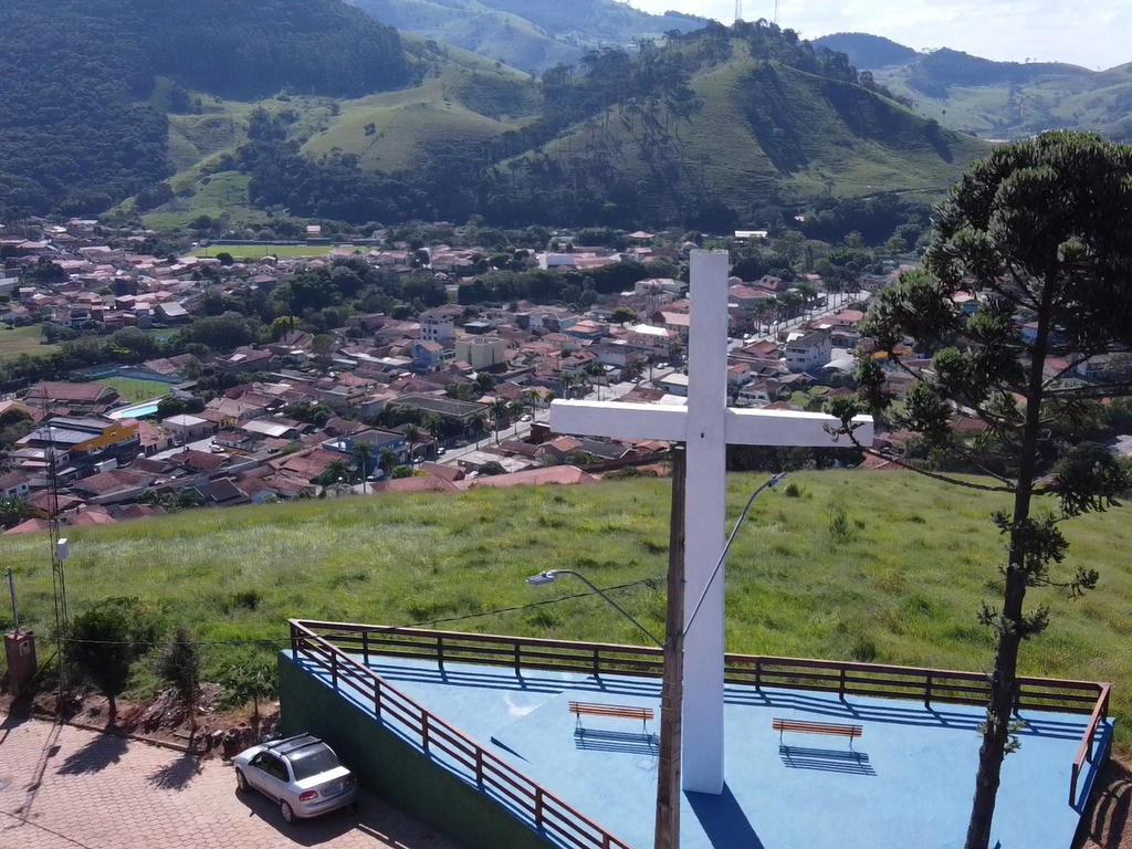 Cruz branca sobre uma plataforma azul com vista para uma cidade e montanhas verdes.
