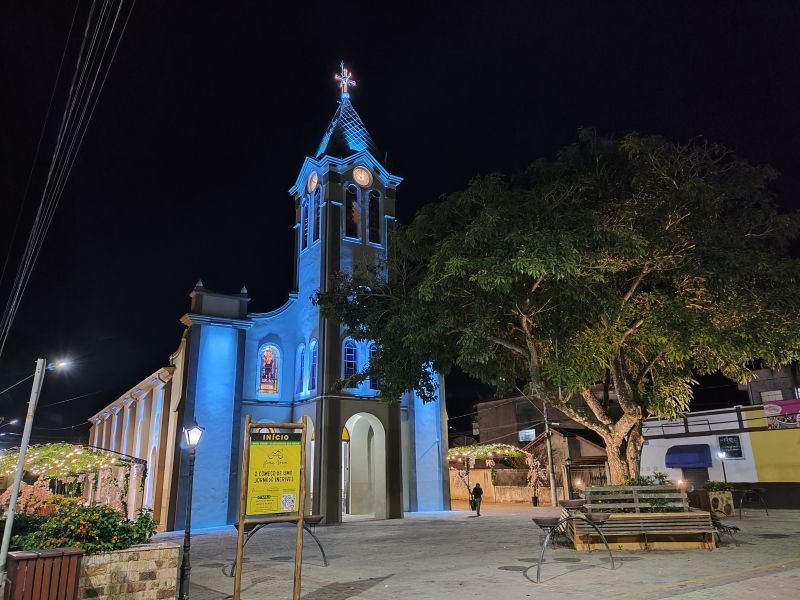 Igreja azul à noite com torre iluminada, entrada em arco e uma grande árvore ao lado de um banco em primeiro plano.