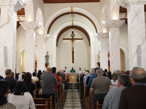 Fiéis de pé no corredor da igreja durante um culto, de frente para o altar e o grande crucifixo.