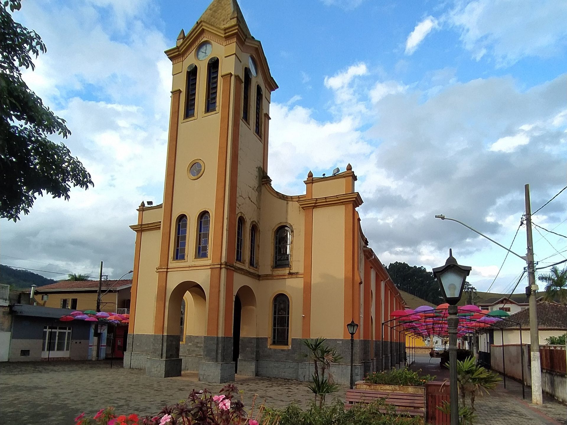 Alta torre amarela de igreja em uma praça sob um céu nublado, com guarda-chuvas coloridos por perto.