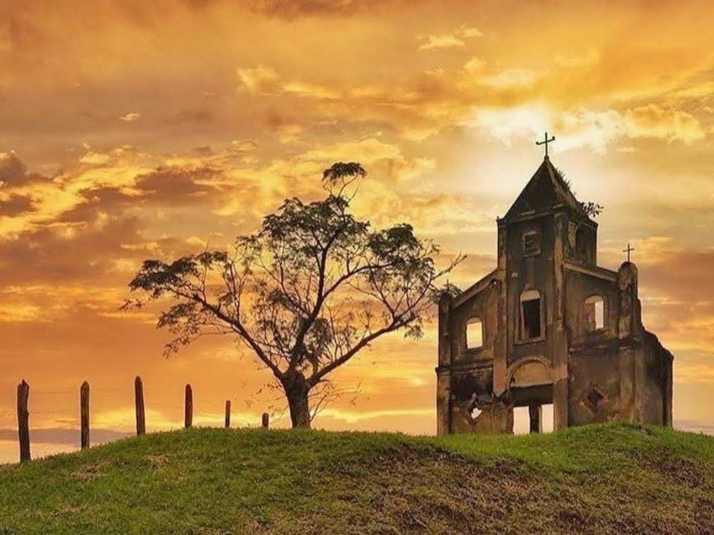 Igreja antiga numa colina ao pôr do sol, ao lado de uma árvore solitária e postes de cerca.