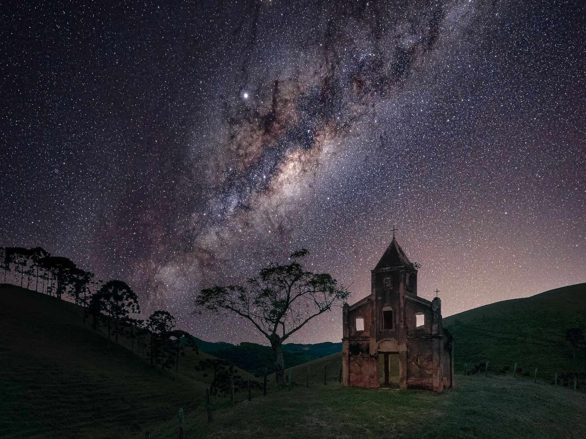 Casa de pedra antiga sob um céu estrelado da Via Láctea ao entardecer, em uma paisagem rural gramada.