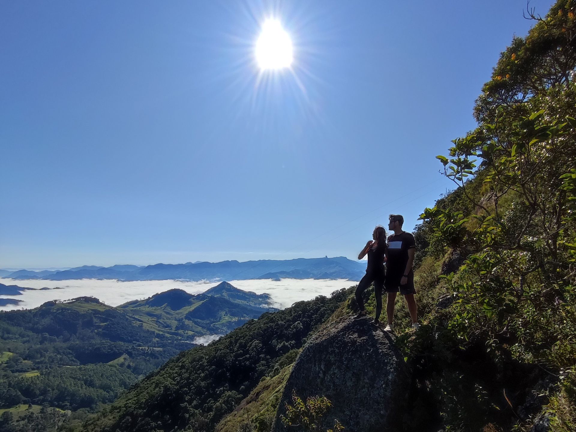Dois excursionistas estão em um cume rochoso acima de montanhas verdes e nuvens sob um céu azul brilhante.