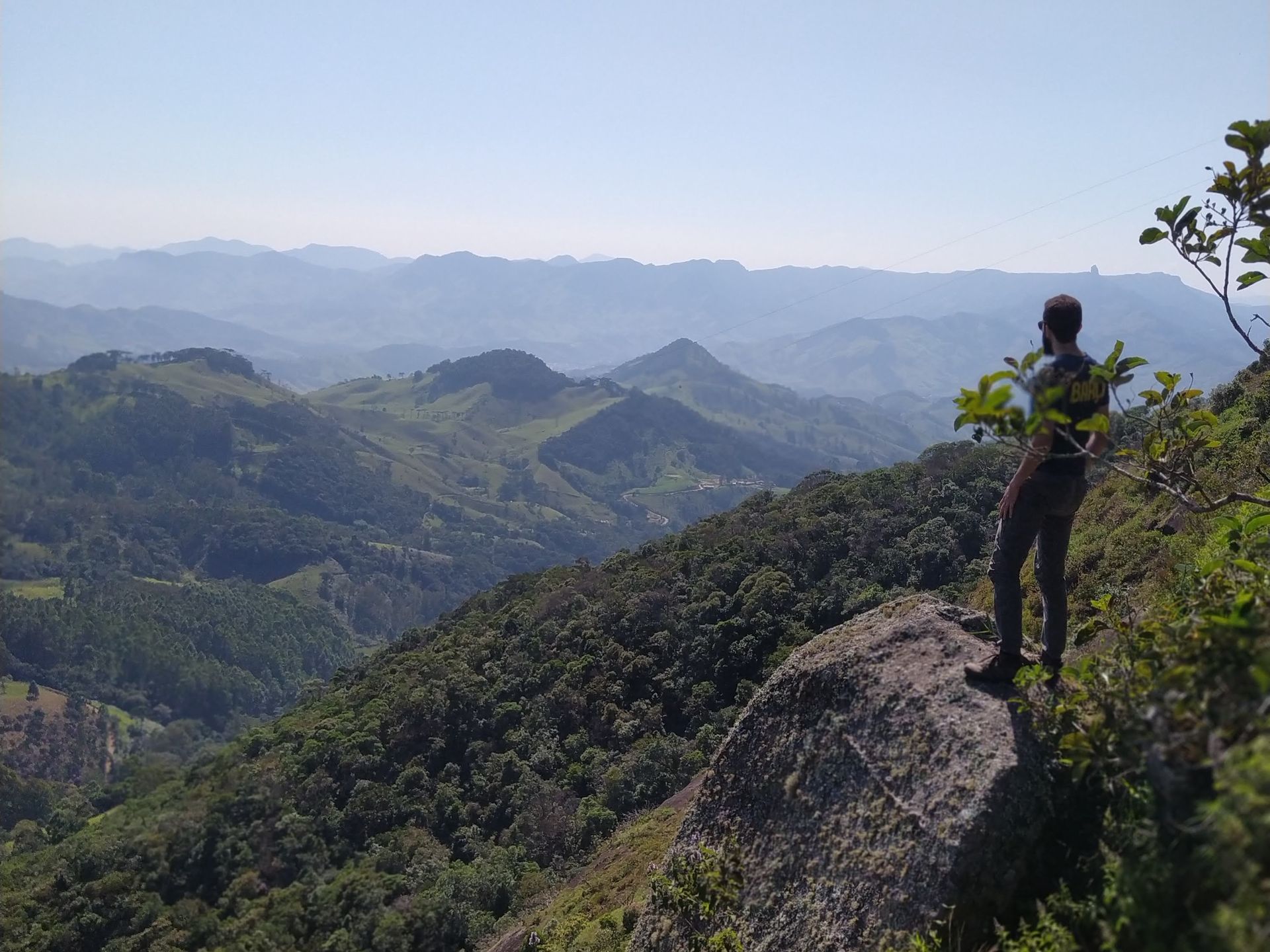 Caminhante em pé em um cume rochoso com vista para vales verdejantes sob um céu azul enevoado.