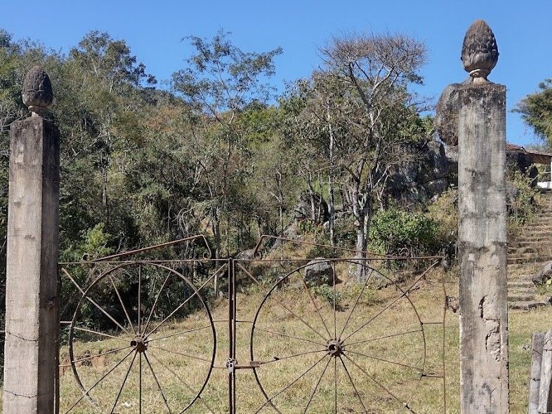 Portão de metal rústico entre postes de pedra em um campo gramado e seco com árvores ao fundo.