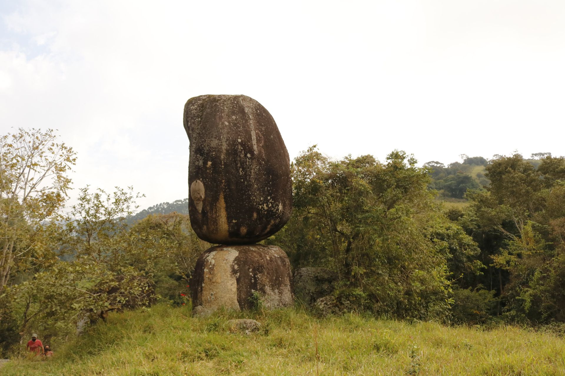 Grande pedra escura equilibrada sobre uma rocha menor em um campo gramado com árvores.