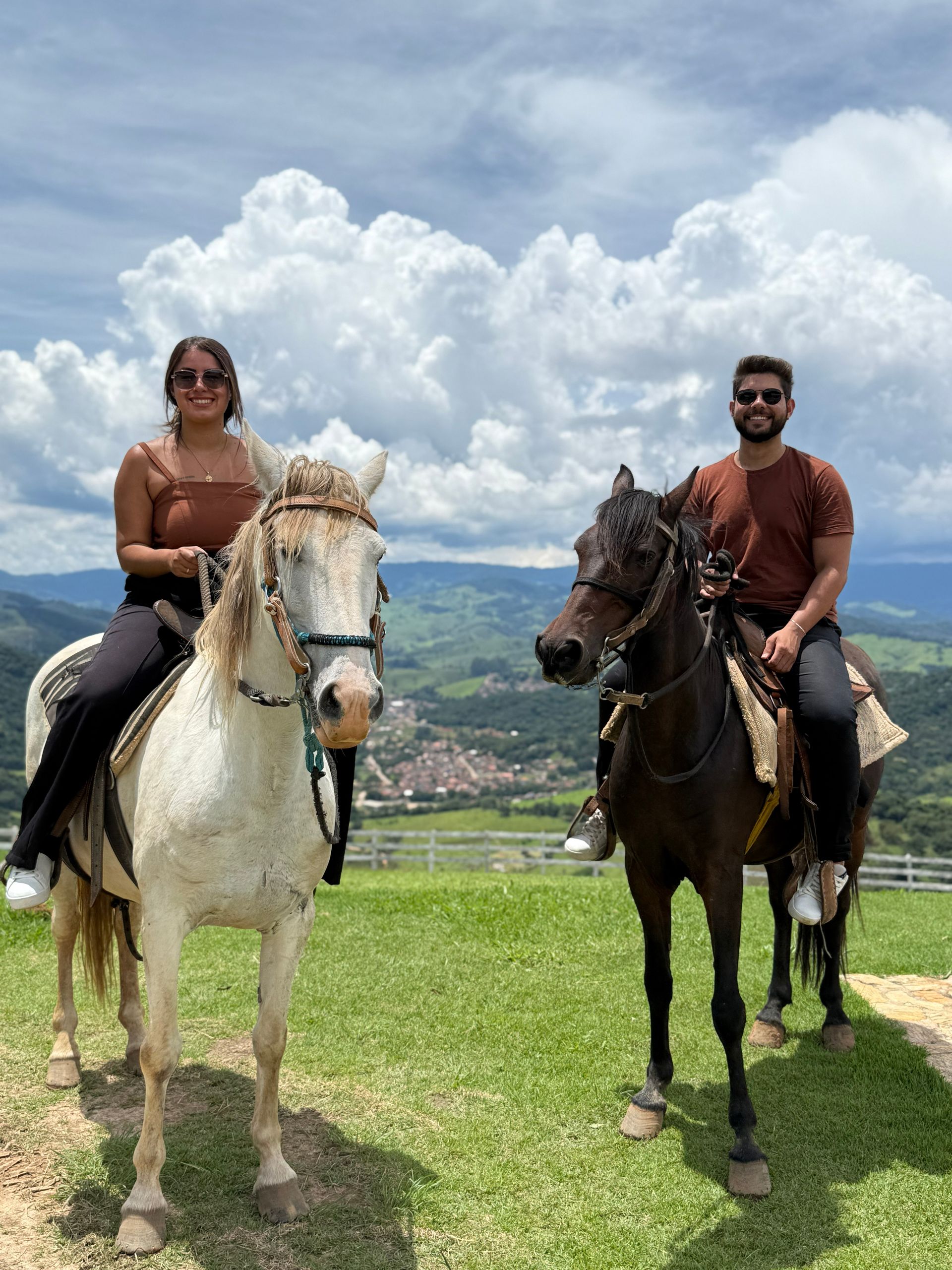 Dois cavaleiros posando em uma colina gramada com montanhas e nuvens ao fundo.