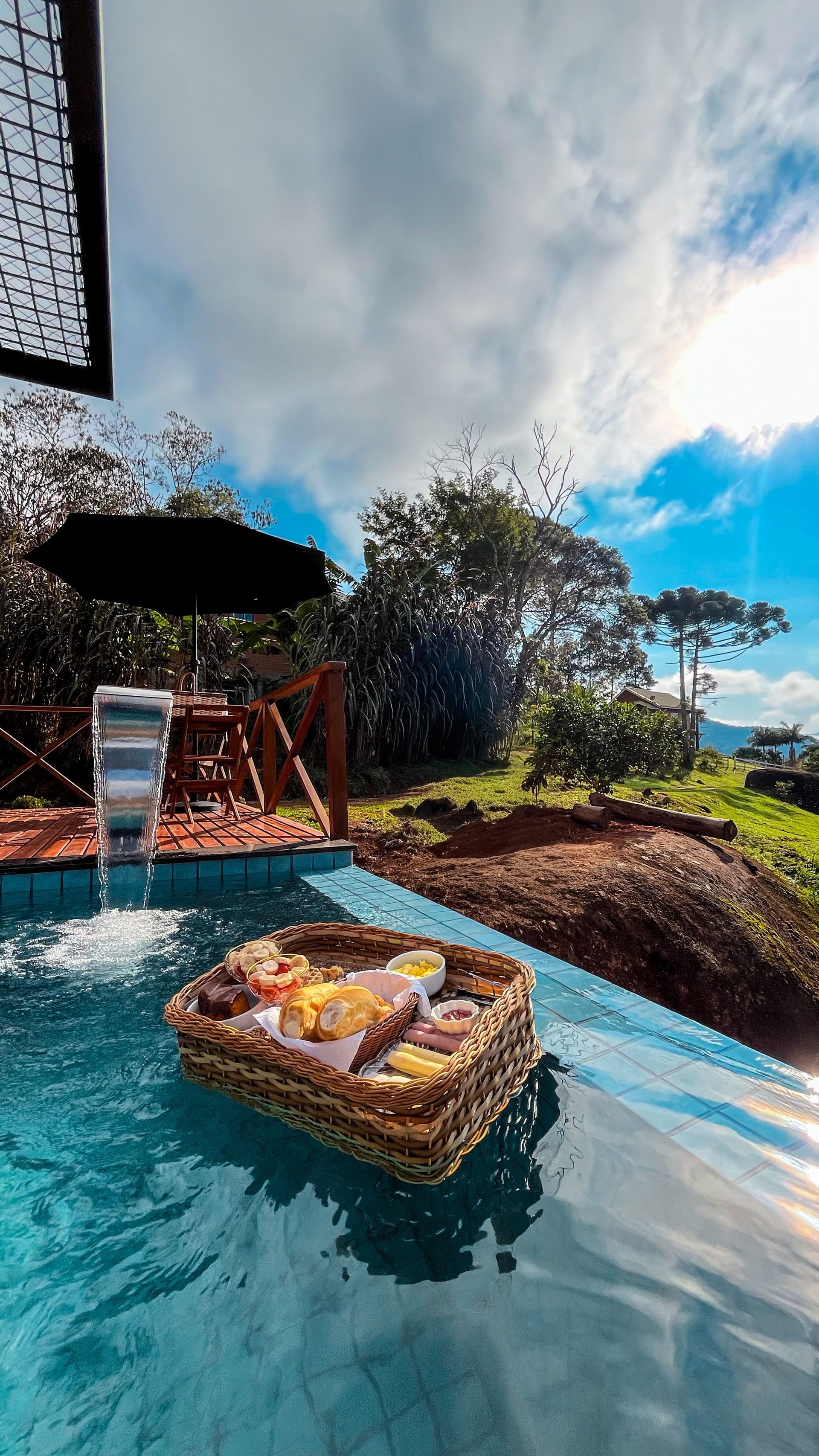 Mesa externa com cesta de vime e bebidas com vista para um jardim ensolarado e céu azul.